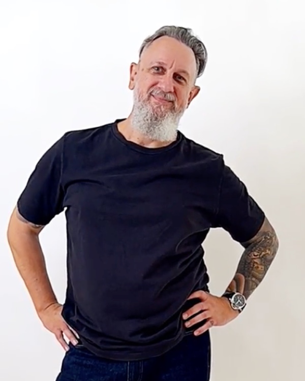 Robert Turrall at 60 — grey-bearded, hands on hips, in a black t-shirt against a plain backdrop